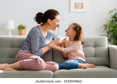 Mom And Her Child Daughter Are Playing At Home. Cute Girl Are Holding Paper Mustache On Stick. Family Holiday And Togetherness.