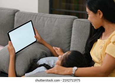 Mom And Her Child Looking On Digital Tablet During Her Daughter Laying On Mom's Lap, Empty Screen Of Tablet, Happy Family Are Spending Time Together, Mother's Day Concept. 