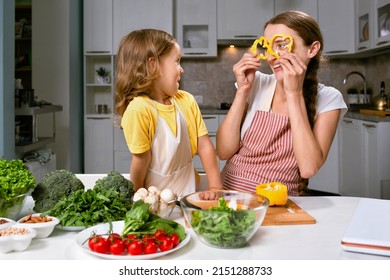 mom and her daughter playfully cook healthy and proper food from fresh vegetables and herbs.The girl makes herself a kind of glasses from pepper circles - Shutterstock ID 2151288733