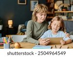 Mom and her daughter with similar haircuts sitting at wooden desk and watching cartoons via tablet together