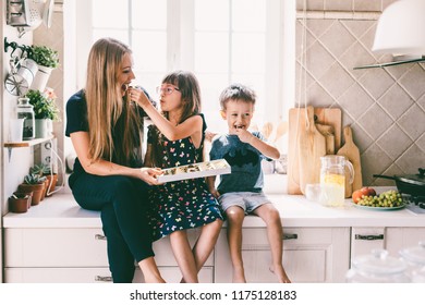 Mom with her two children sitting on the kitchen table and eating candies. Mother with daughter and toddler son having breakfast at home. Happy lifestyle family moments.