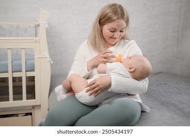 Mom holds baby in arms, sitting on bed next to crib, and feeds newborn baby with formula from bottle, showing concern for child. Health care, newborn feeding concept, alternative to breastfeeding.  - Powered by Shutterstock