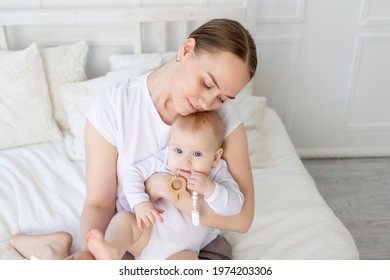 Mom Holds Baby In Her Arms With A Rodent On A White Bed With Cotton Bedding At Home, Teether