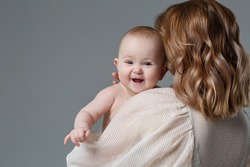 Mom Holds A Cute Baby In Her Arms, View Over Her Shoulder, Isolated On A Gray Background.