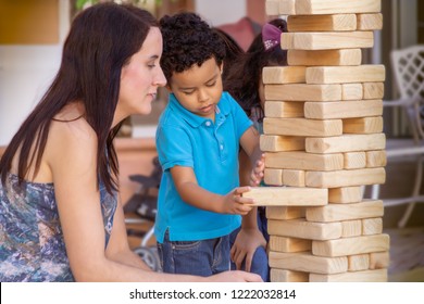 Mom Holds Her Breath As Her Little Boy Carefully Removes A Block From The Jumbling Wood Tower. The Wooden Large Size Jumbling Tower  Holds Steady While The Boy Keeps It In Place With The Other Hand. 