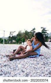 Mom Hugs A Little Girl, Bending Over Her While Sitting On The Beach