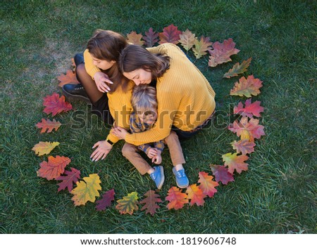 Mom hugs  two her children. family sits next to each other on  grass. Around them is a circle of colorful autumn leaves. Family joy, tenderness, happy motherhood. top view