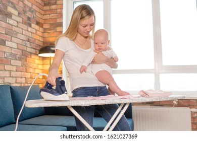 Mom Irons Children's Clothes With An Iron On An Ironing Board Holding A Newborn Baby In Her Arms. Mom's Household Chores