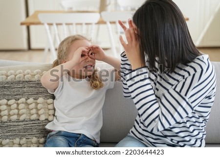 Mom and kid little daughter makes funny faces, having fun, playing together, sitting on sofa at home. Mother and small girl showing glasses by hands, looking to each other, fooling around.