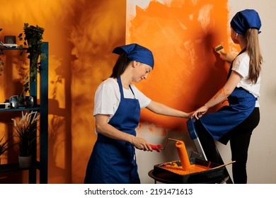 Mom And Kid Renovating House With Paint, Using Orange Color To Decorate Wall Surface In Apartment. Young Child On Ladder Painting With Paintbrush And Adult With Rolling Brush, Having Fun.