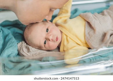 Mom kisses her newborn baby who is resting in a transparent cradle in the hospital. This tender moment emphasizes the bond between mother and child, creating a cozy and safe atmosphere for the infant - Powered by Shutterstock