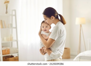 Mom Kissing Baby. Happy Young Woman Standing In Bedroom Or Nursery Room Interior In Tender Pastel Colors Holding And Kissing Cute Sweet Beautiful Little Child. Family, Love, Care, Mother's Day Concept