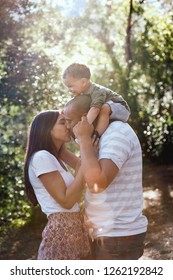 Mom Kissing Dad With Kid On His Shoulders, Happy Family Outdoors In Forest