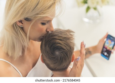 Mom Kissing Her Little Son On The Head And Watching Something In The Phone With Him. High Quality Photo