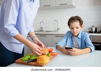 Mom Is In The Kitchen Preparing A Homemade Burger For Her Teenage Son.
An 11-year-old Boy Is Sitting At The Kitchen Table And Looking At His Mobile Phone.