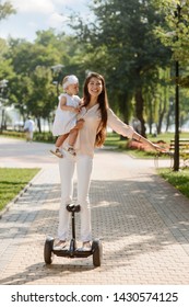 Mom And Little Daughter Taking A Fast Ride On An Electronic Scooter. Fun And Amusement In The City Park.