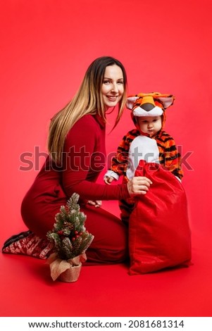 Mom with a little son with a small Christmas tree and a red bag with gifts. Beautiful smiling brunette in a burgundy dress and a child in a tiger fur suit - the simbol of 2022. Red background. 