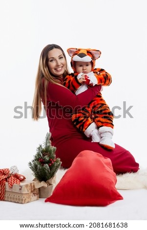 Mom with a little son with a small Christmas tree and a red bag with gifts. A beautiful brunette in a burgundy dress and a child in a tiger fur suit - the simbol of 2022. White background. Vertical.