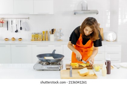 Mom on orange apron standing beside flying pan and gas stove in modern home kitchen learning to cook tasty family food for kid by half cutting flied breads.