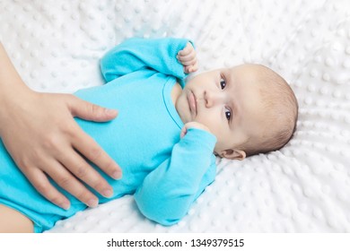 Mom Pats The Tummy Of A Small Child, Mom Takes Care Of Her Son, Baby In Blue Pajamas Lying On A White Blanket