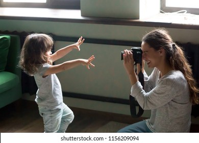 Mom photographer having fun playing making photo of artistic cute kid daughter at home, young mother photographing little child girl or shooting video on digital camera, funny family photo session