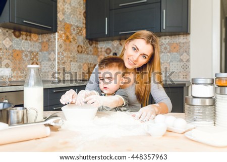 Mom playing with kid in the kitchen. Kitchen is done is dark colors and roustic style. Kid is covered in flour and looks funny. Table is made from light wood. Blender, milk, eggs, jars stand on table.