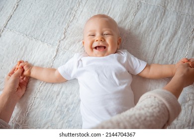 Mom Plays With The Baby, Gives Him A Massage, Teaches Him To Sit Down And Control His Body. Child In A White Bodysuit On A White Bed. Healthy Active Kid. Lifestyle