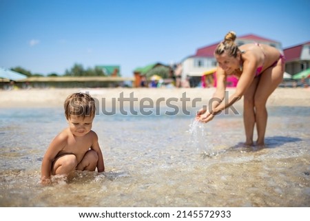 Mom plays and splashes on her baby who is laughing while standing in the sea under the summer sun on vacation