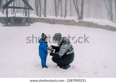 Mom puts on mittens to a little child squatting in a snowy park