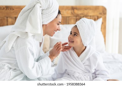 Mom Putting Make Up On Her Cute Little Daughter, Side View, Closeup. Cheerful Mother In Bathrobe With Hair Wrapped In Towel Applying Lipstick On Her Kid Lips, Bedroom Interior