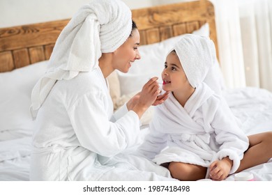 Mom Putting Make Up On Her Cute Little Daughter, Side View, Closeup. Cheerful Mother In Bathrobe With Hair Wrapped In Towel Applying Lipstick On Her Kid Lips, Bedroom Interior