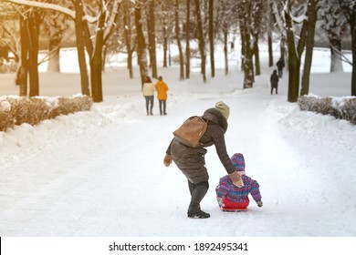 Mom Rides Her Daughter On Board From Hill In Winter Park. Little Children Sliding From Hill At City Park. Winter Snow Sliding. Happy Mother Pushes Daughter On Her Snow Board As She Learns To Slide