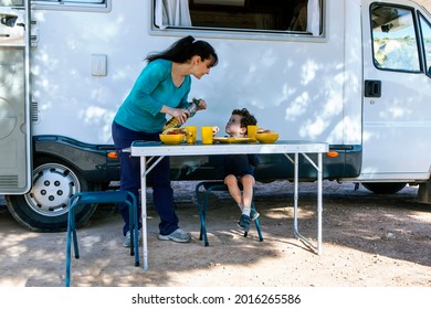 Mom Serving Food To Her Son In Front Of The Motor-home, At The Table In The Shade Of The Tree.