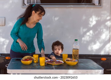 Mom Serving Food To Her Son In Front Of The Motor-home, At The Table In The Shade Of The Tree.