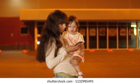 Mom With Small Baby Dials Phone Number On Touchscreen Phone In Night City Under Light Of Street Lamps.