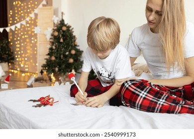 mom and son 7 years old in Christmas pajamas, sitting on bed, write letter to Santa Claus or postcard for friends, dreaming about gifts. Merry Christmas . preparation for winter holidays. Wish list - Powered by Shutterstock