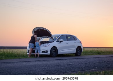 Mom With Son Looking At The Camera While Opening The Front Of A Defective Car At The Grassy Ground.