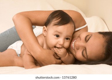 Mom And Son Lying Down On Bed And Mother Embracing The Infant Baby, Who Put His Hand Into Mouth