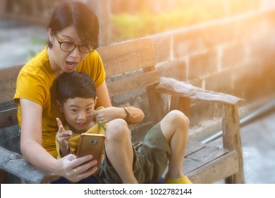 Mom And Son Selfie With Mobile Phone On Wooden Armchair. Asian Black Hair 40s Woman And 4 Year Old Boy Happiness To Using Smart Phone Taking Photos With Sunset Effect. Selective Focus And Copy Space