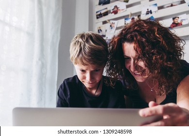 Mom Son Watch Video Computer Smiling. Curly Woman With Blond Child Have Fun Playing With Student Pc. Looking Homework Information Helped By Mother At Home. Family Domestic Scene White Modern Furniture