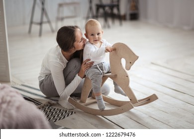 Mom Supports Baby Son Swinging On A Rocking Toy Horse. Real Light Interior, Caring Motherhood Concept