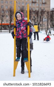 Mom Supports From The Back Of The Child Standing On Wooden Stilts At Winter. Social Activities Are At Maslenitsa Holiday, Russia