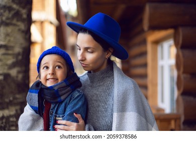 Mom In A Sweater And A Blue Hat With A Child Sitting On The Porch Of A Wooden House In Autumn