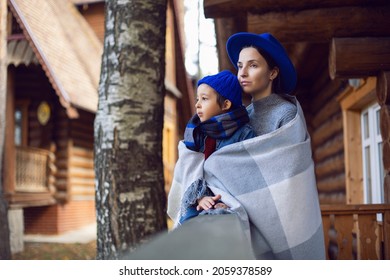 Mom In A Sweater And A Blue Hat With A Child Sitting On The Porch Of A Wooden House In Autumn