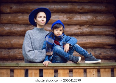 Mom In A Sweater And A Blue Hat With A Child Sitting On The Porch Of A Wooden House In Autumn