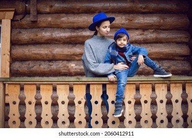 Mom In A Sweater And A Blue Hat With A Child Sitting On The Porch Of A Wooden House In Autumn