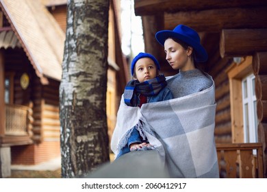 Mom In A Sweater And A Blue Hat With A Child Sitting On The Porch Of A Wooden House In Autumn