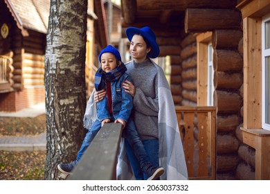 Mom In A Sweater And A Blue Hat With A Child Sitting On The Porch Of A Wooden House In Autumn