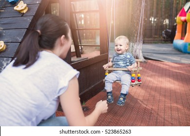 Mom Swinging Baby Boy In Swing At Summer Time. Toddler With Mother Having Fun Outdoors In The Park. Happy Family Lifestyle Concept