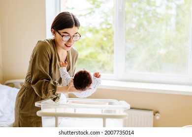 Mom Takes The Newborn Baby From The Crib In Postpartum Ward. . Mother Kissing And Touching Baby With Love And Care.